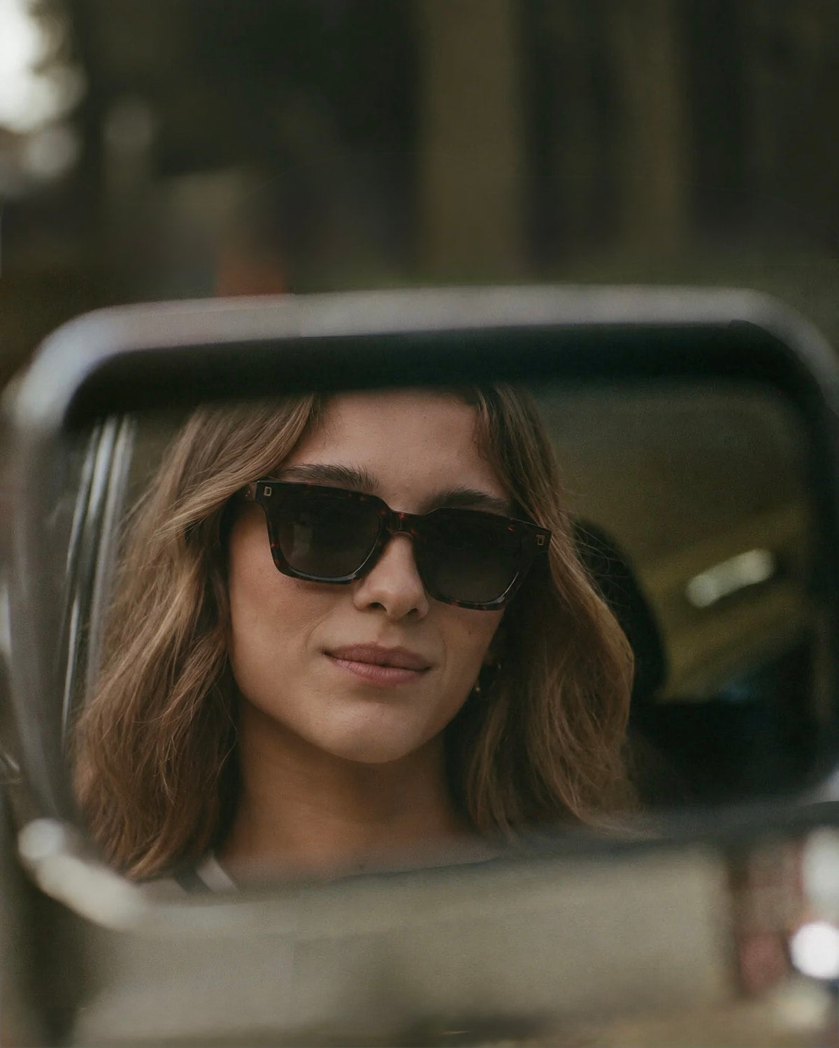 Una mujer con lentes de sol de acetato se ve reflejada en el espejo lateral de una camioneta.