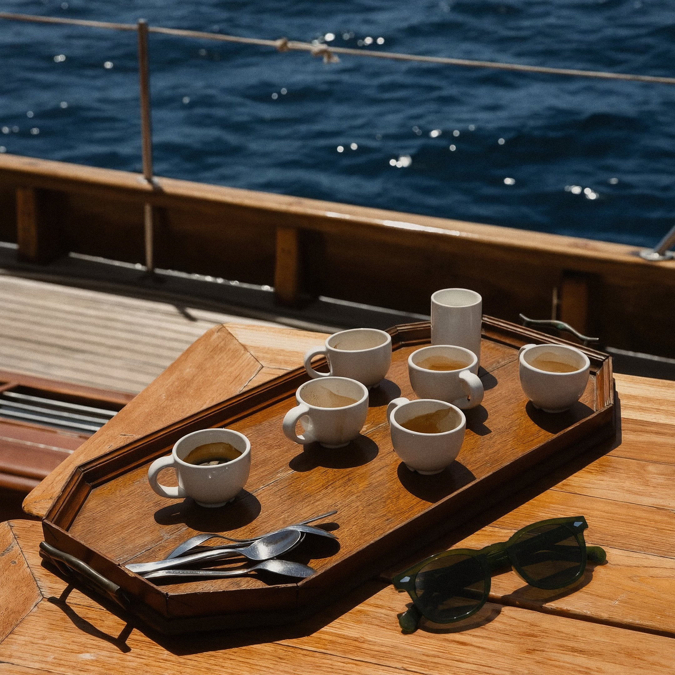 Una bandeja de madera con siete tazas de café y cucharas sobre la cubierta de un velero, junto a unos lentes de sol verdes, con el mar azul brillando al fondo.