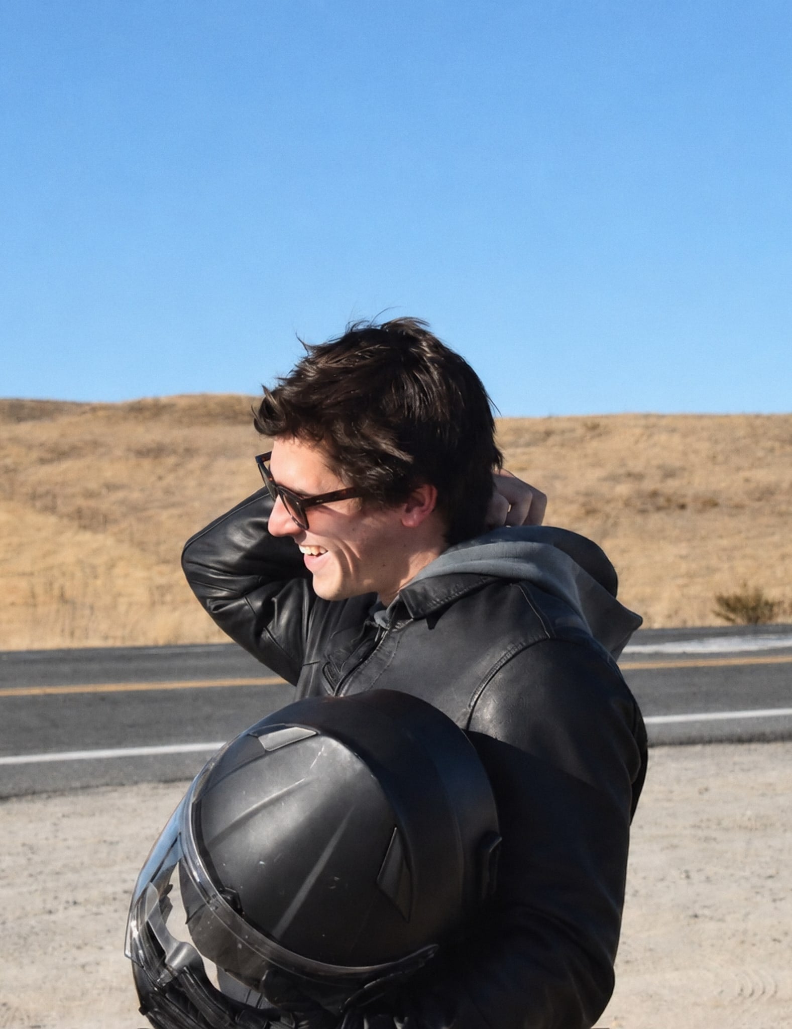 Alguien riéndose a la orilla de una carretera con el casco de moto en los brazos.