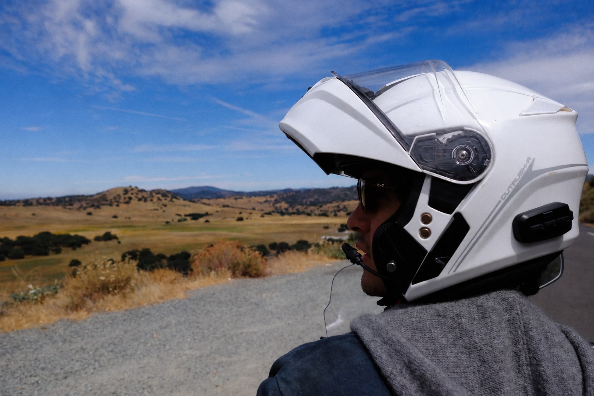 Alguien con casco de moto blanco mirando un valle de cerros secos desde la orilla de una carretera.