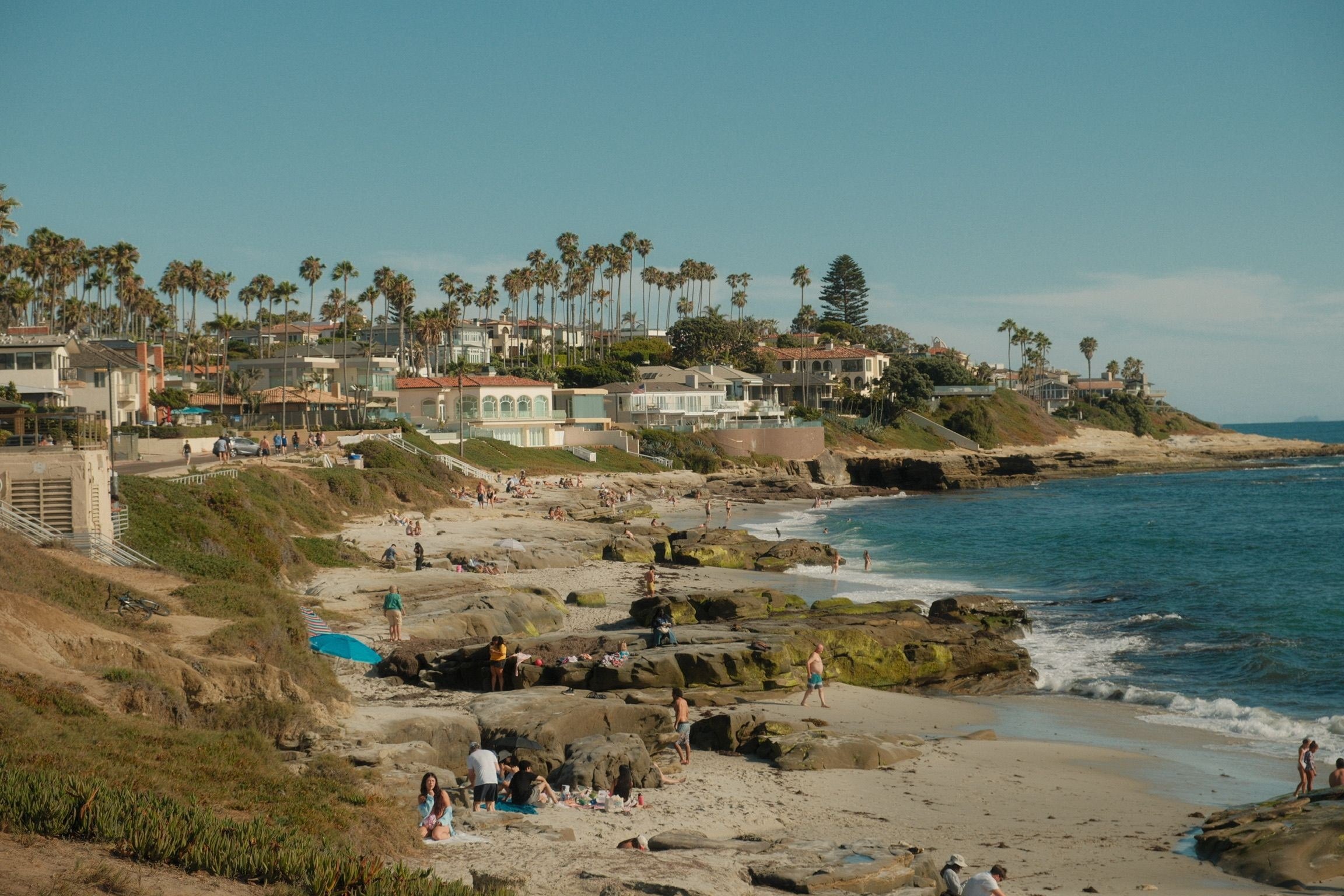 Una playa con rocas y gente al sol, y en el acantilado una fila de casas entre palmeras.