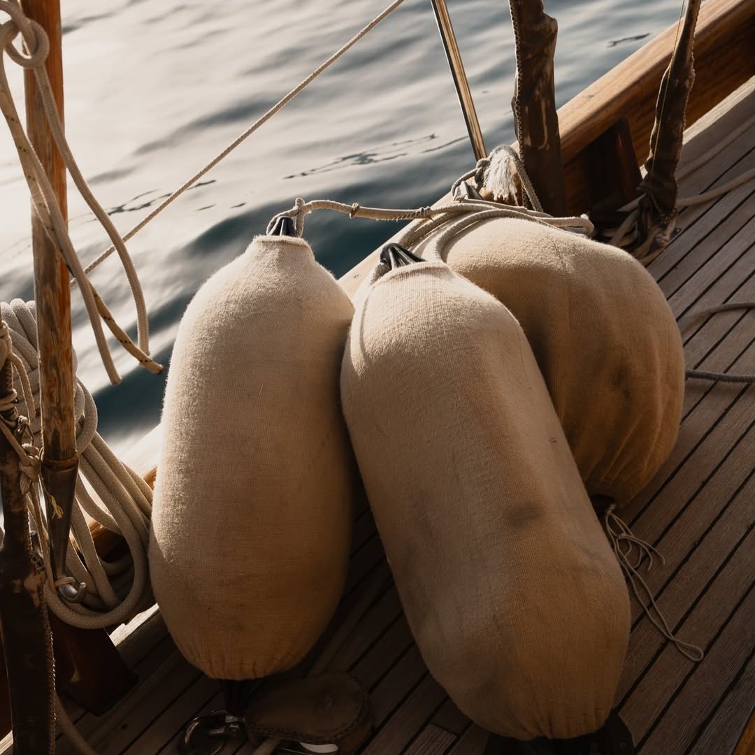 Tres defensas de lona beige amarradas sobre la cubierta de madera de un velero, con cabos, un candelero y el mar al fondo con luz baja.