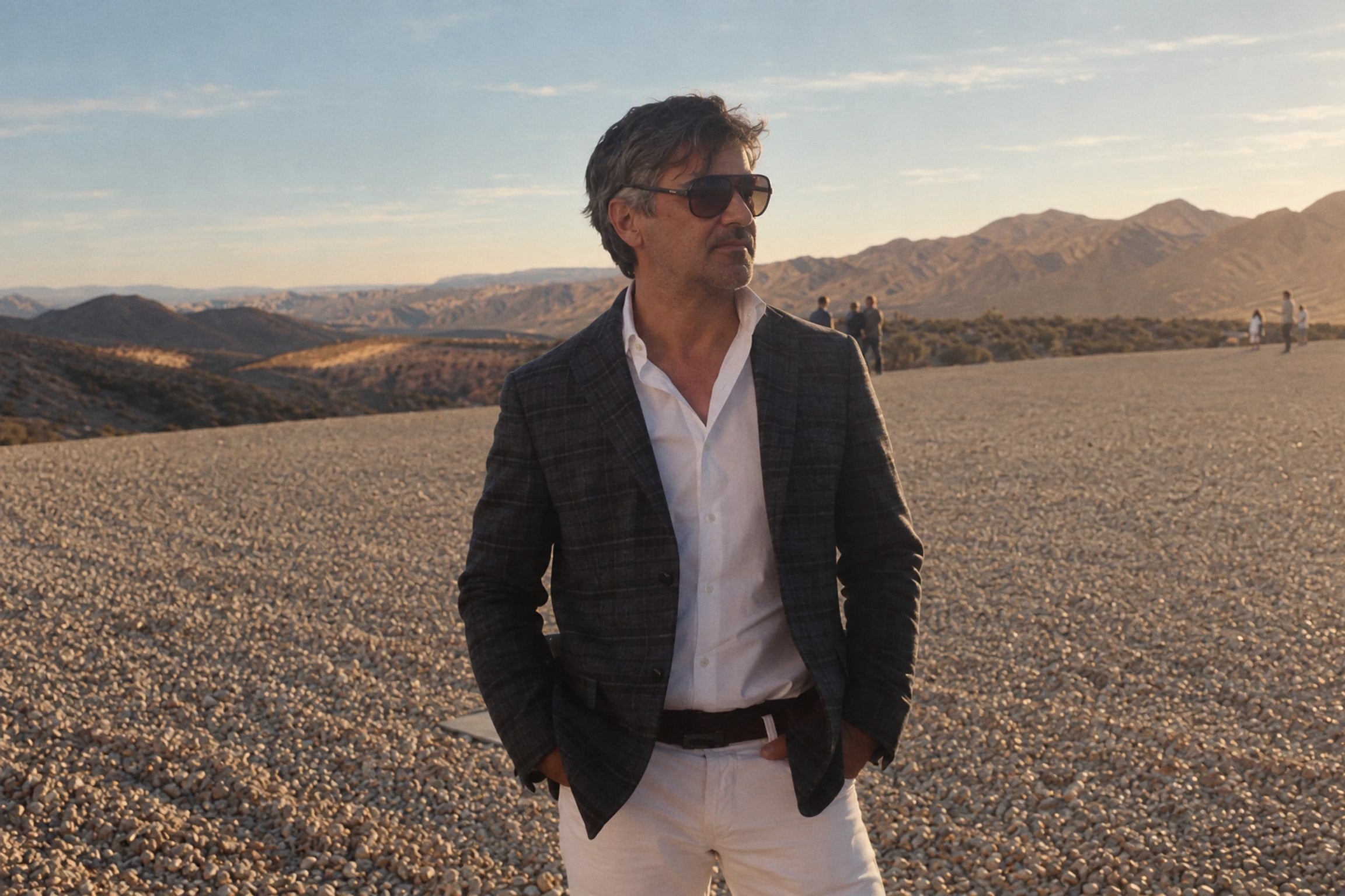 Alguien de pie en un llano de grava con lentes de sol, saco y camisa blanca, con montañas detrás al atardecer.