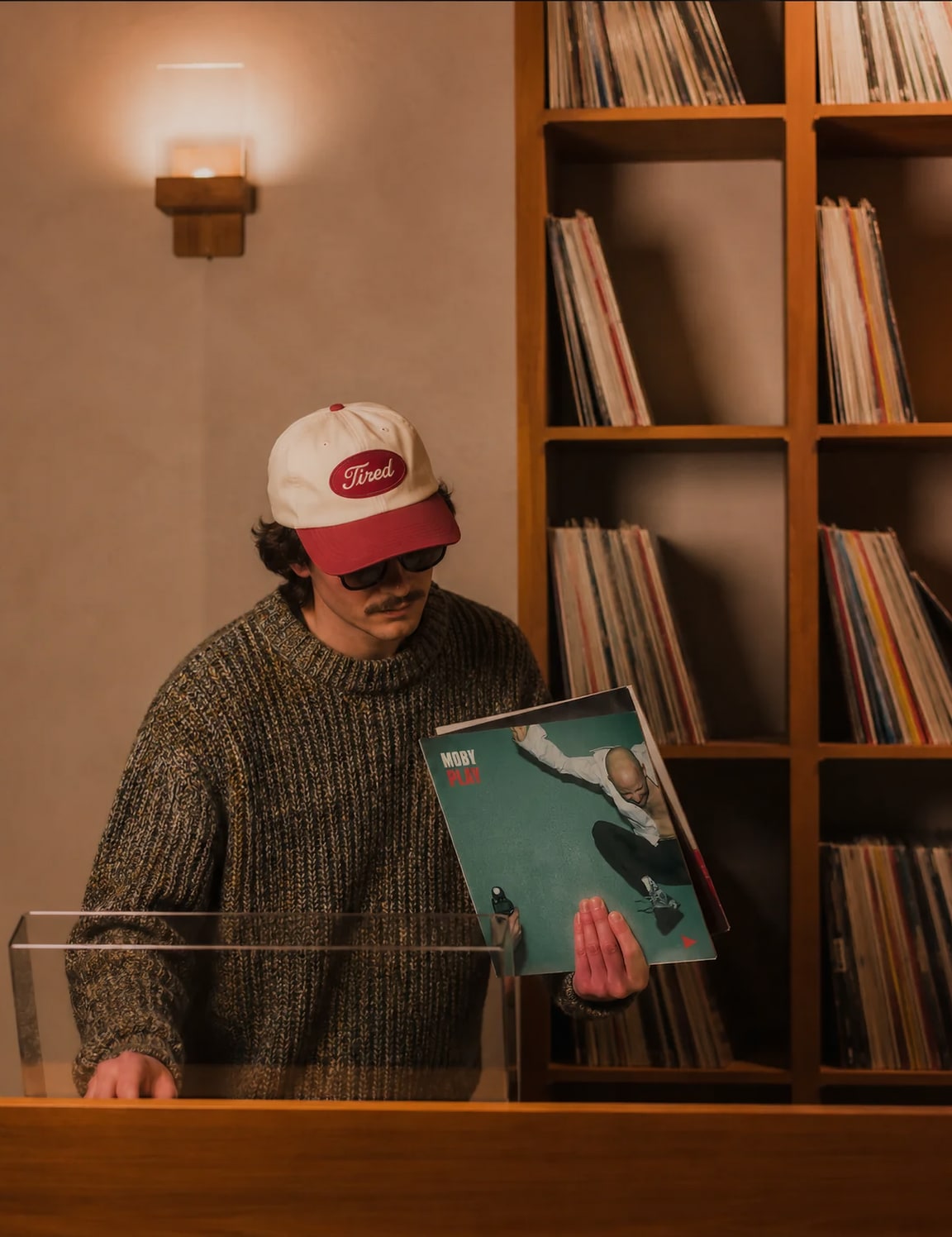 Un hombre con gorra revisa un disco en una tienda de vinilos, frente a una estantería de madera llena de discos.