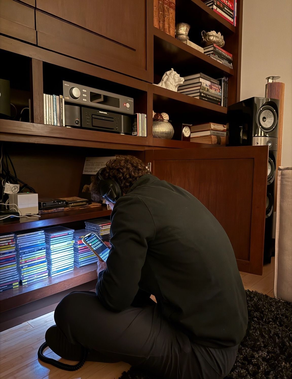 Alguien en cuclillas frente a un mueble con equipo de sonido, discos y libros, revisando música en el teléfono.