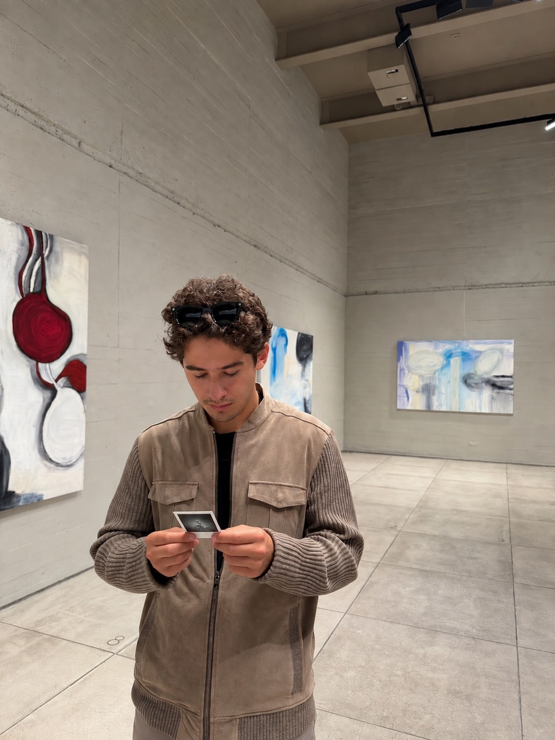 Un hombre de pie en una sala de concreto con cuadros abstractos, mirando algo que sostiene con las dos manos.