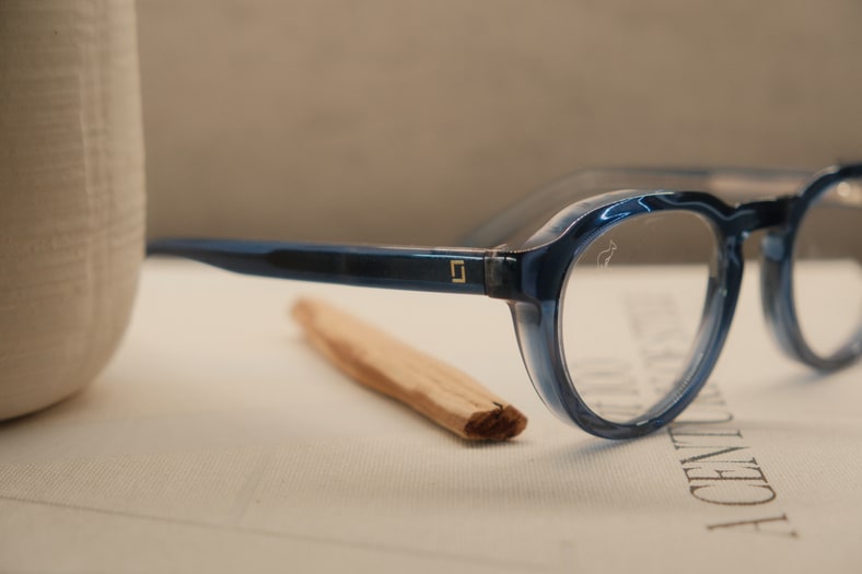 Unos lentes de acetato azul apoyados sobre un libro, junto a un jarrón de barro y un palo santo.
