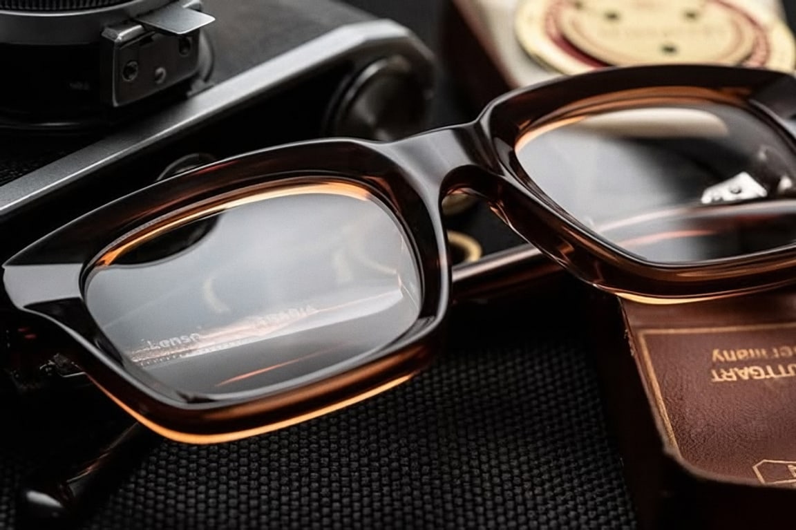 Primer plano de unos lentes de acetato ámbar sobre una superficie oscura, con una cámara y un estuche de piel al lado.