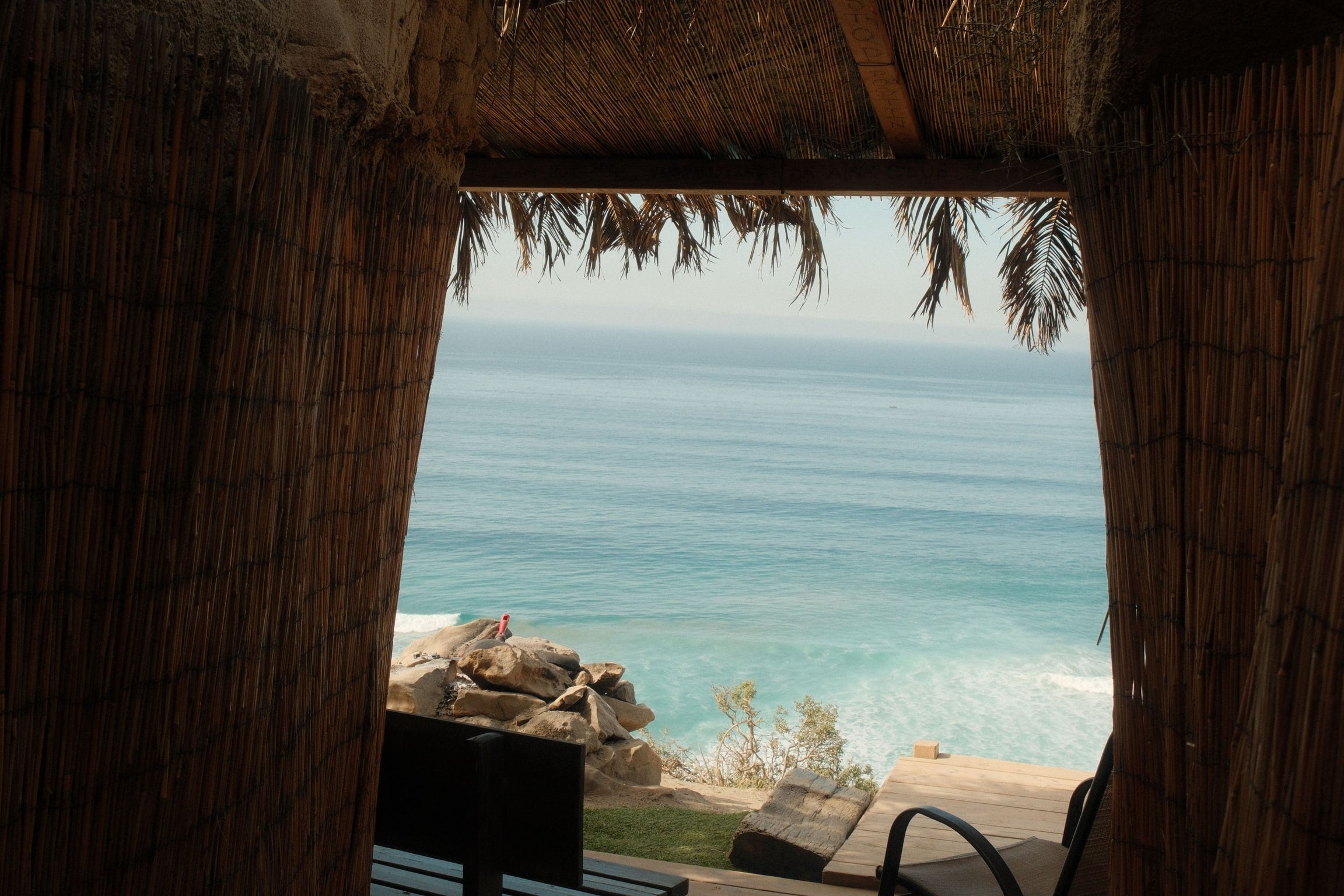 Vista desde el interior de una palapa de carrizo hacia el mar, con la terraza de madera y las rocas al fondo.