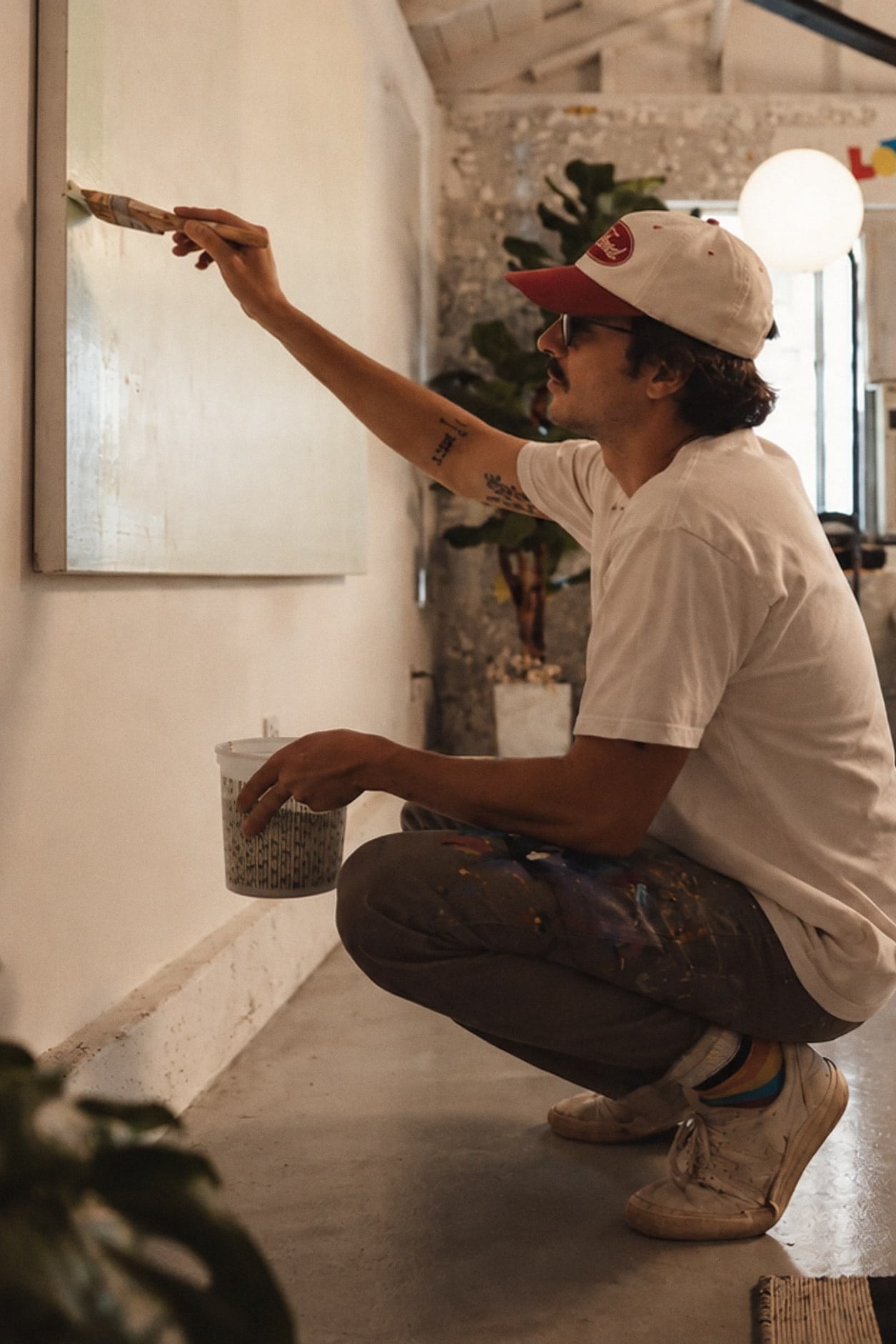 Alguien en cuclillas pintando un cuadro colgado en la pared de un taller con mucha luz.