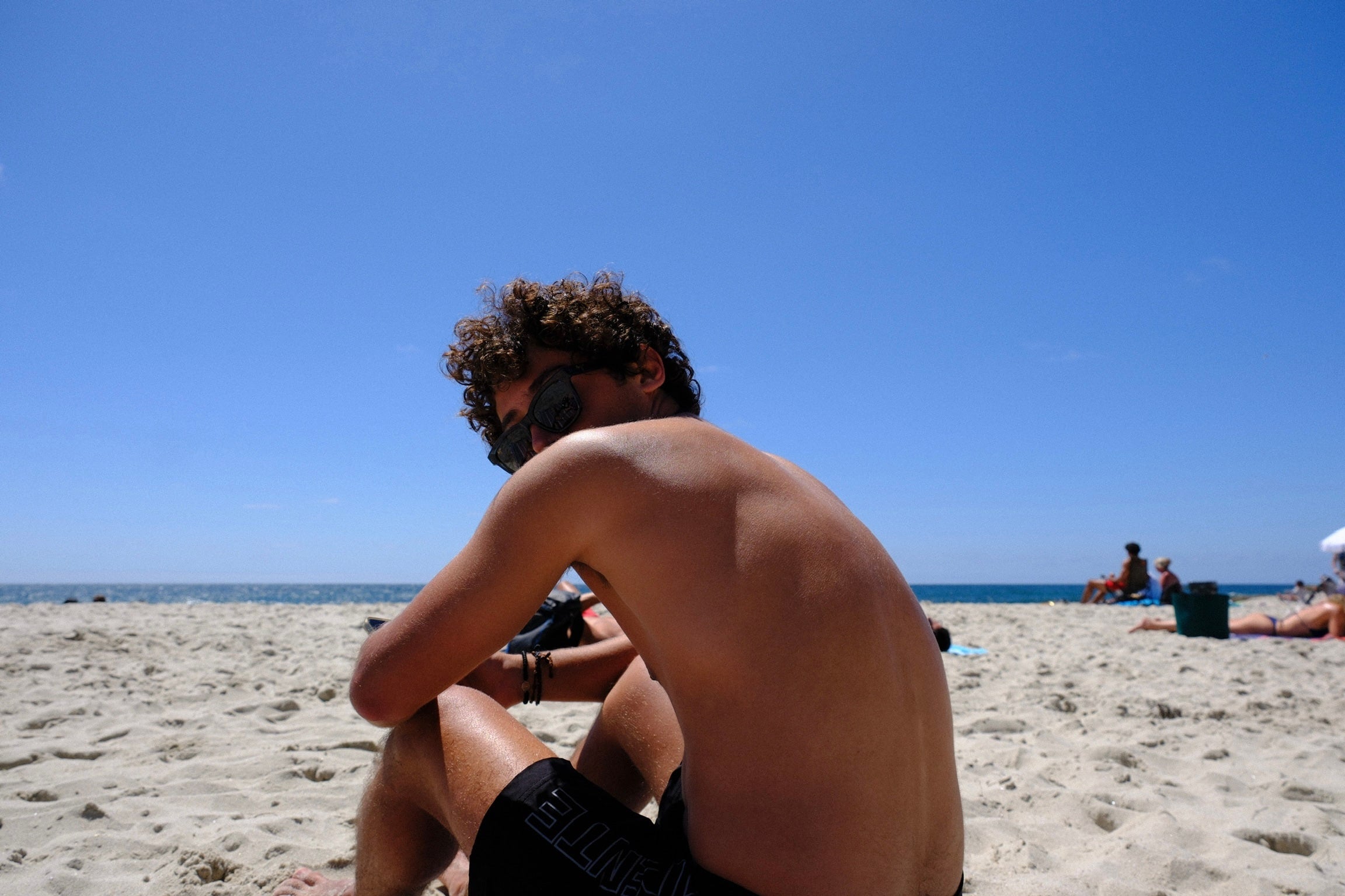 Alguien sentado en la arena de una playa con un visor de buceo puesto, de espaldas al mar.