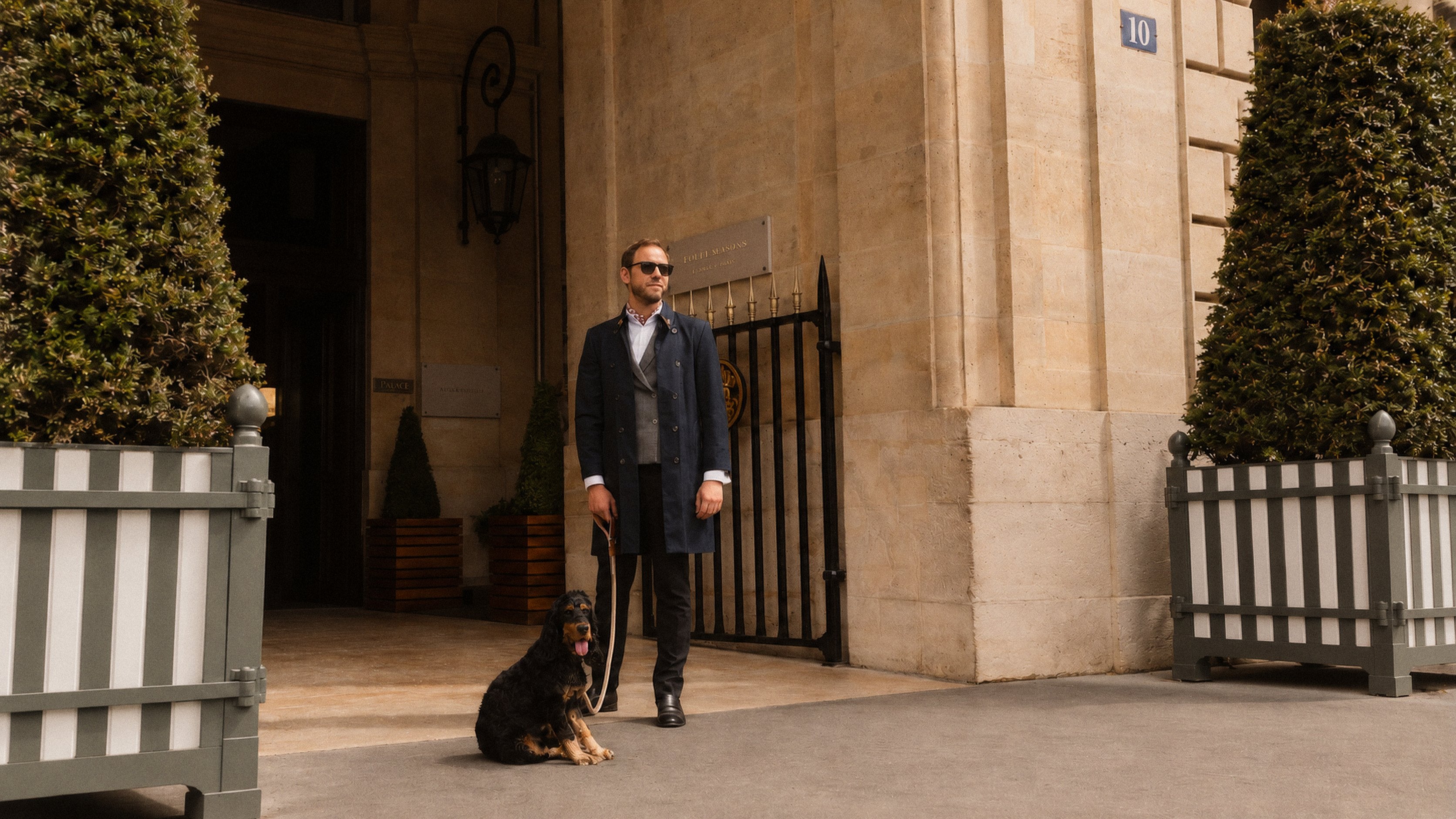 Un hombre con lentes de sol y abrigo azul marino, de pie frente al portal de piedra de un edificio, con un perro negro sentado a su lado y macetas con arbustos recortados a ambos lados.
