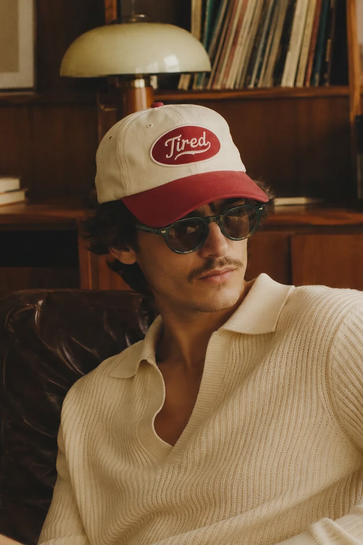Un hombre con gorra y lentes de sol sentado en un sillón de piel, frente a un mueble con discos y un amplificador.