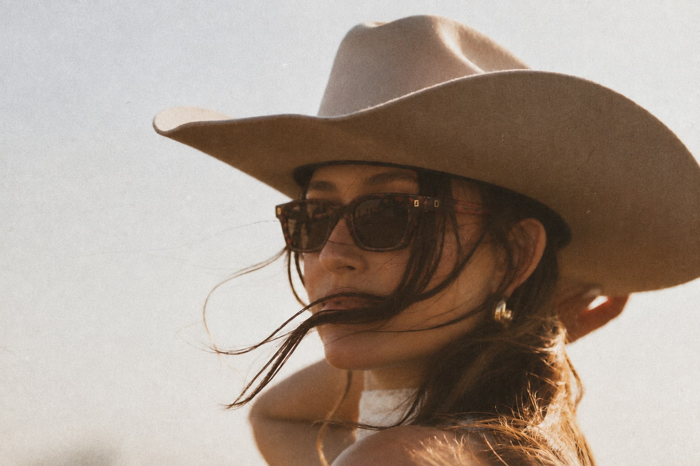 Alguien con sombrero vaquero claro y lentes de sol, a contraluz, con el pelo cruzándole la cara por el viento.