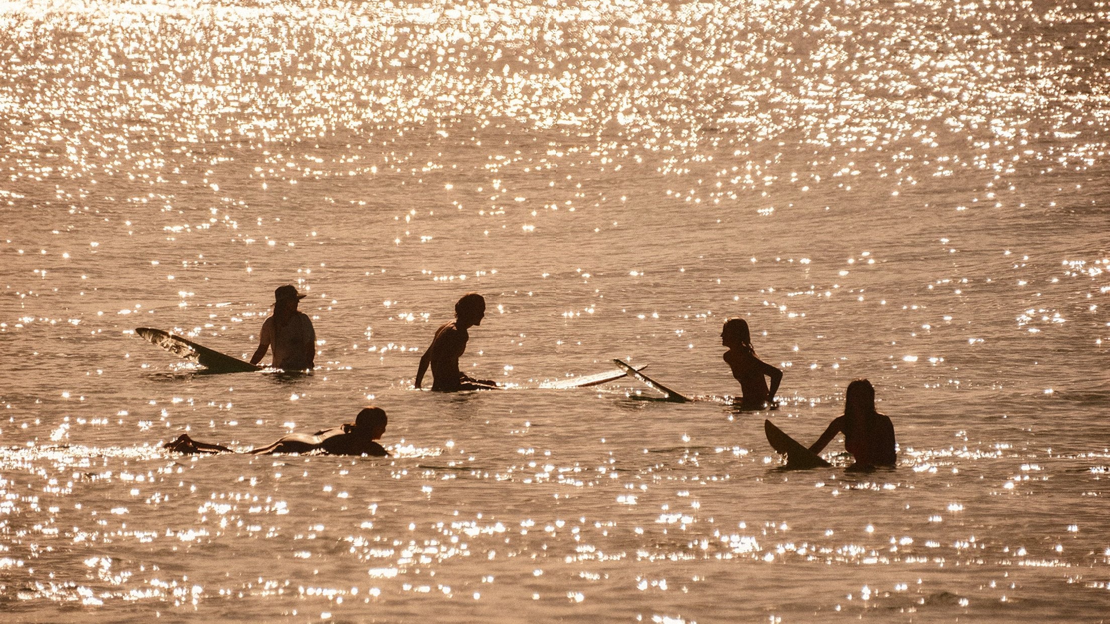 Cinco surfistas sentados en sus tablas, a contraluz, esperando ola sobre un mar que brilla dorado con el sol bajo.