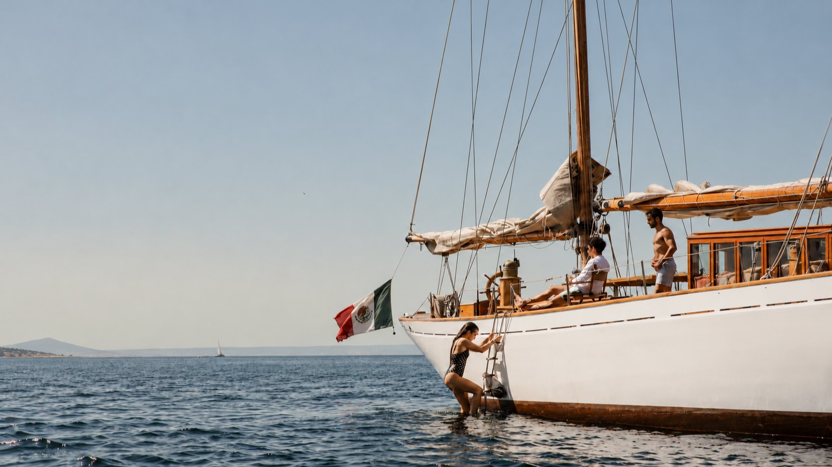 Un velero de madera con bandera de México fondeado en mar tranquilo; una persona baja por la escalera del casco y dos más descansan en cubierta.