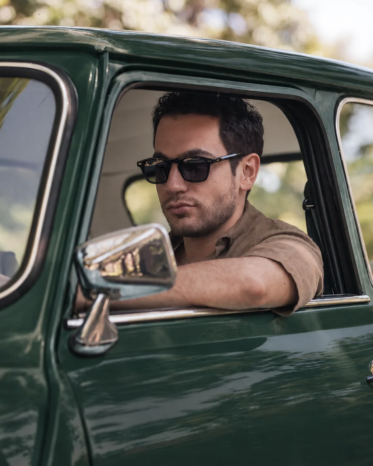 Un hombre con lentes de sol asoma por la ventanilla de una camioneta verde, con el brazo apoyado en la puerta.